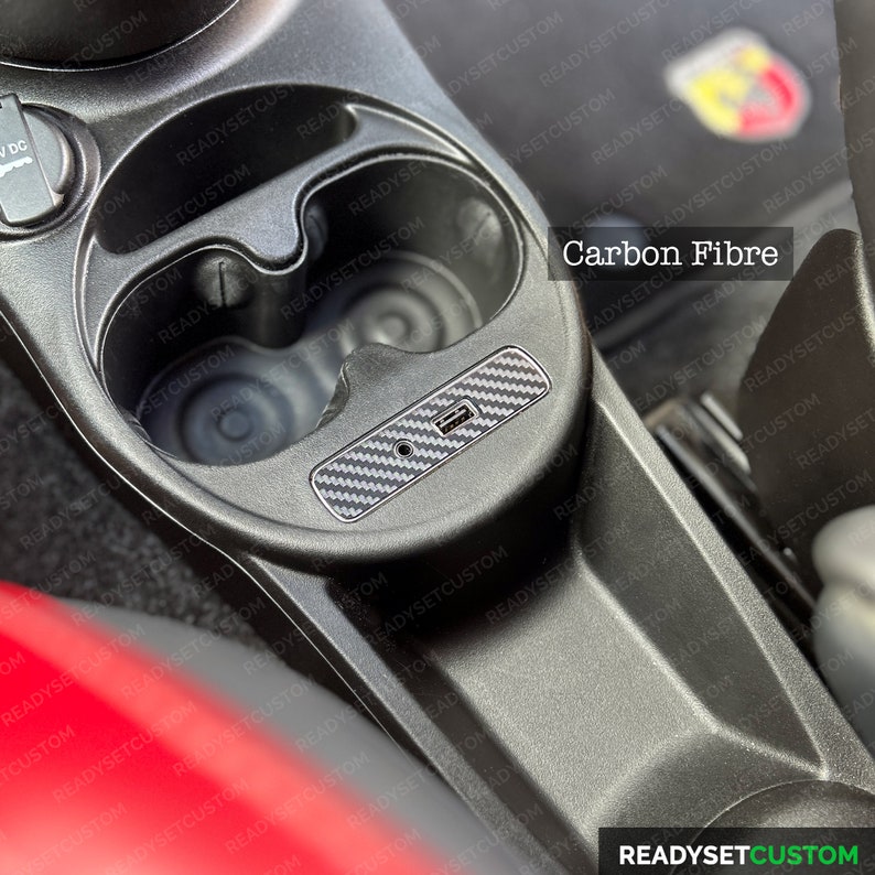May include: A close-up of a car's interior with two cup holders and a carbon fiber trim piece. The trim piece has a USB port and the text "Carbon Fibre" is visible.