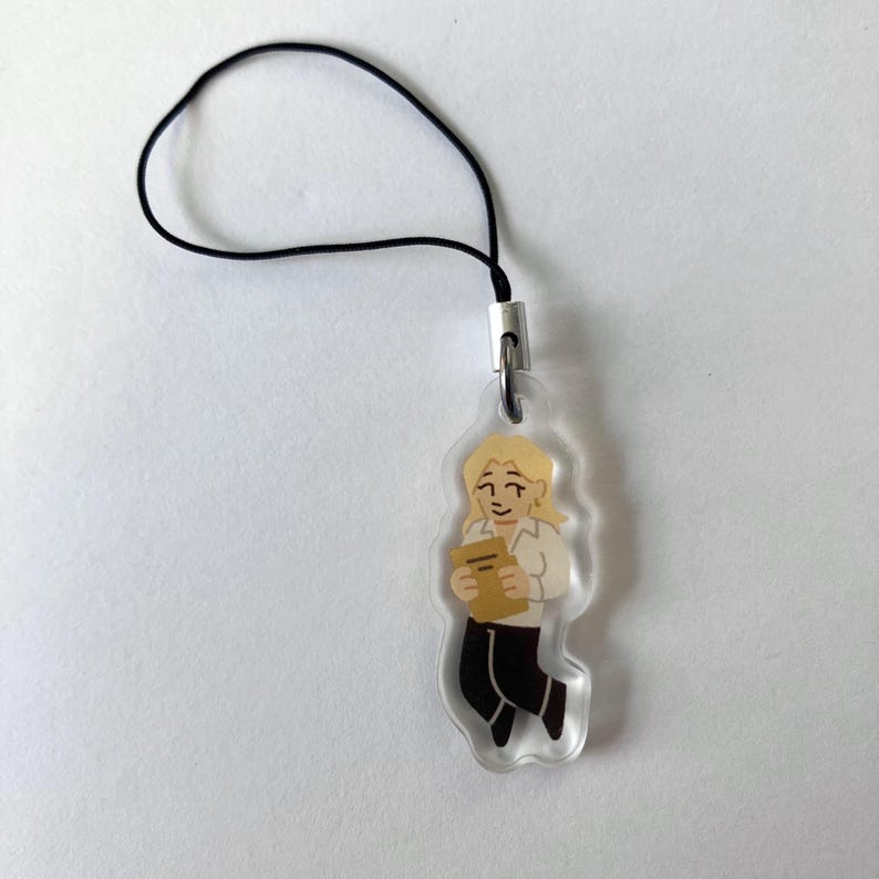 May include: A clear acrylic charm featuring a cartoon illustration of a person with blonde hair holding a book. The charm has a black cord loop for attachment. The person is wearing a white shirt and black pants.