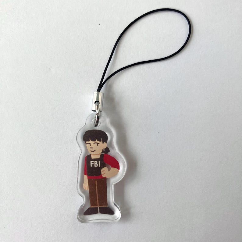 May include: Clear acrylic charm featuring a cartoon illustration of a person in an FBI vest and red shirt. The charm has a black cord for attaching to a phone or bag. The word "FBI" is printed on the vest.