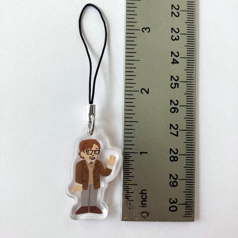 May include: A clear acrylic charm featuring a cartoon illustration of a person in a brown jacket, glasses, and gray pants. The charm is approximately 1 inch in height and has a black cord attached for hanging.