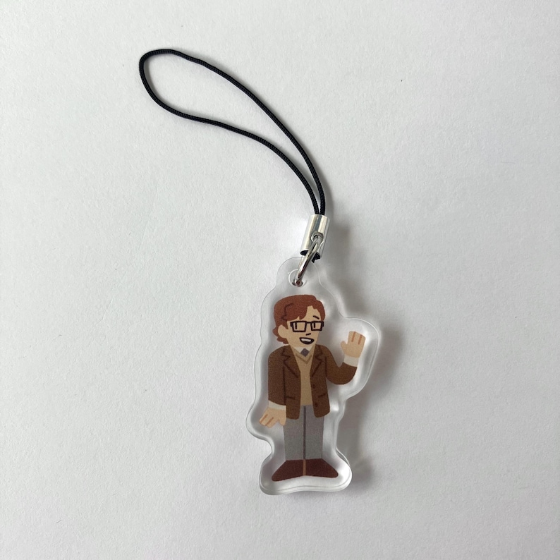 May include: A clear acrylic charm featuring a cartoon illustration of a man with brown hair, glasses, and a brown jacket. The charm has a black lanyard attached for easy hanging. The man is waving with his right hand. The charm is a fun accessory.