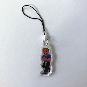 May include: A clear acrylic charm featuring a cartoon illustration of a person with brown skin, wearing a blue shirt and black pants. The charm has a black cord and silver-colored metal clasp for attachment. The person is smiling with their arms crossed.