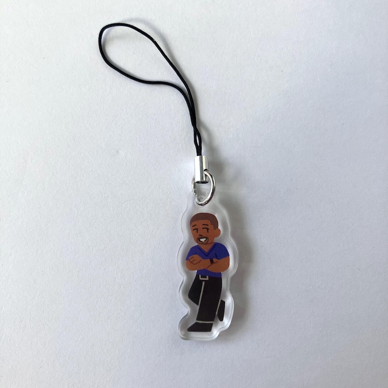 May include: A clear acrylic charm featuring a cartoon illustration of a person with brown skin, wearing a blue shirt and black pants. The charm has a black cord and silver-colored metal clasp for attachment. The person is smiling with their arms crossed.