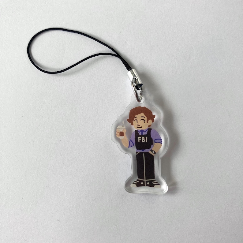 May include: Clear acrylic charm featuring a cartoon illustration of a person in an FBI vest holding a coffee cup. The charm has a black lanyard attached for hanging. The FBI logo is visible on the vest.