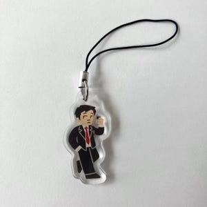 May include: Clear acrylic charm featuring a cartoon figure in a black suit and red tie, holding a small object. The charm has a black cord and silver-tone metal clasp for attachment. The figure is in a walking pose.