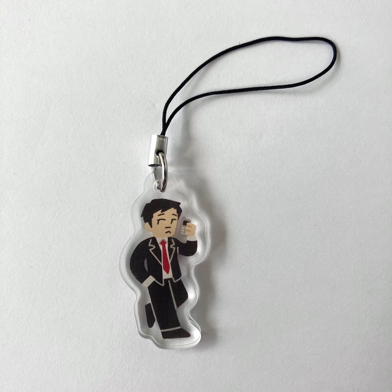 May include: Clear acrylic charm featuring a cartoon figure in a black suit and red tie, holding a small object. The charm has a black cord and silver-tone metal clasp for attachment. The figure is in a walking pose.
