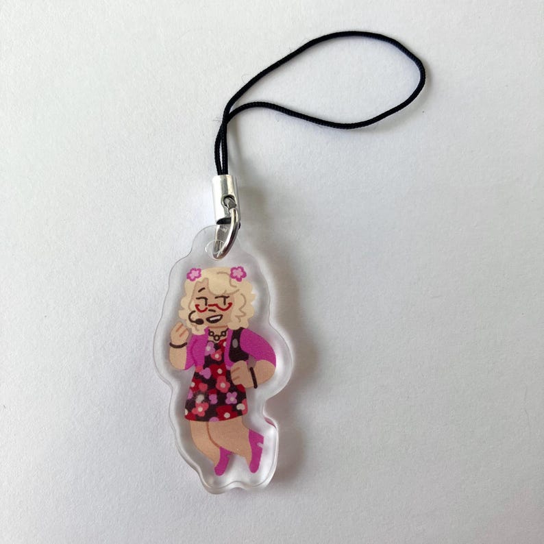 May include: A clear acrylic charm featuring a cartoon character with blonde curly hair, glasses, and a pink outfit with a floral pattern. The charm has a black lanyard attached for hanging. The character is smiling and appears to be holding a microphone.