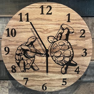 May include: A round wooden clock with a light brown finish. The clock face features black numerals and two black turtle illustrations. One turtle plays a banjo, and the other dances. The clock hands are black.