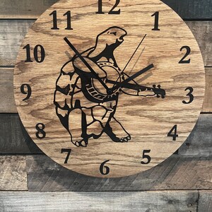 May include: A round wooden clock with a light brown finish. The clock face features black numerals and hands. A black silhouette of a turtle playing a banjo is in the center. The clock is mounted on a wooden wall.