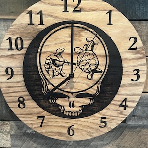 May include: Wooden wall clock with a light brown finish and black numerals. The clock face features a black graphic of turtles playing instruments within a skull design. The clock hands are black.