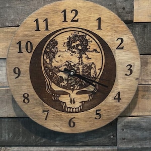 May include: Round wooden clock with a light brown face and dark brown numbers. The clock features a central graphic of a skeleton with roses and a skull. The clock hands are black.