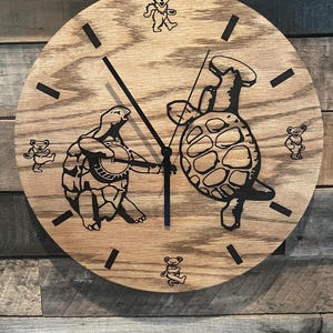 May include: Wooden wall clock with a light brown finish. The clock face features black illustrations of turtles playing musical instruments and dancing bears. Black lines mark the hours. The clock hands are black.