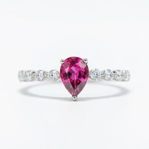 May include: A silver ring featuring a pear-shaped, vibrant pink gemstone as the focal point. The band is adorned with a series of small, round, clear gemstones. The ring is set against a white background, highlighting its elegant design.