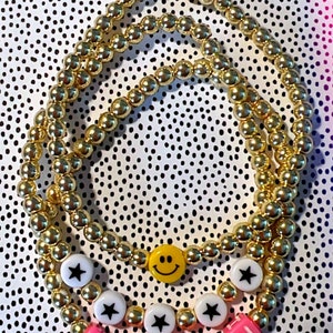May include: Three gold bead bracelets with charms. The charms include a smiley face, a star, and the word "TEACH" spelled out in pink letter beads.