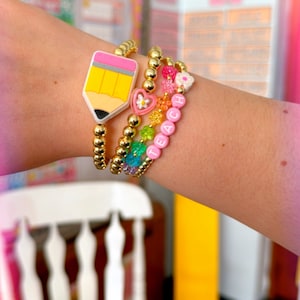 Pode incluir: Um conjunto de quatro pulseiras com contas douradas, um amuleto de lápis amarelo, pingentes de flores rosa, contas de arco-íris e uma pulseira de contas rosa com a palavra "TEACH" escrita.