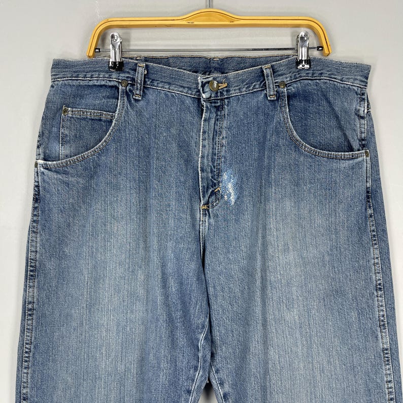 May include: Light blue denim jeans with a classic five-pocket design. The jeans are hanging on a wooden hanger. The jeans have a faded wash and a button and zipper closure.