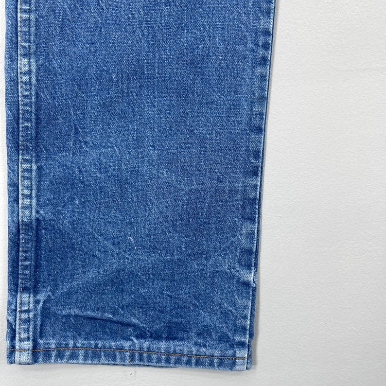 Pode incluir: Close-up de uma se&ccedil;&atilde;o de jeans azul. Os jeans t&ecirc;m um design cl&aacute;ssico de cinco bolsos com costuras vis&iacute;veis e uma lavagem desbotada. A imagem mostra a bainha e a costura lateral dos jeans.