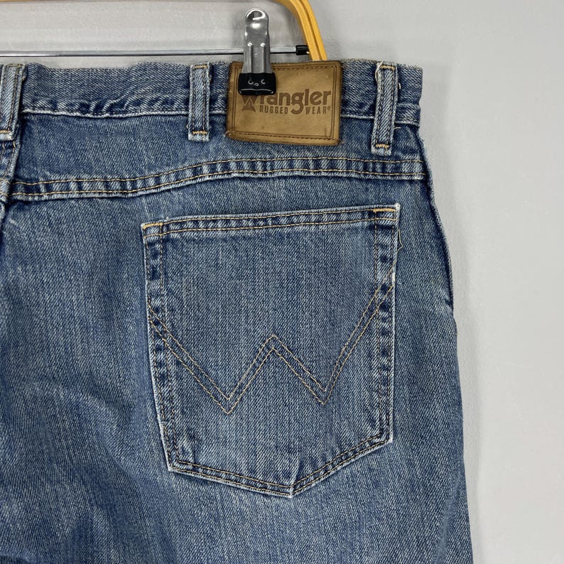 May include: Close-up of blue denim jeans with a back pocket featuring a stitched 'W' design. The jeans have a brown leather patch with the 'Wrangler Rugged Wear' logo. The jeans are hanging from a metal clip.