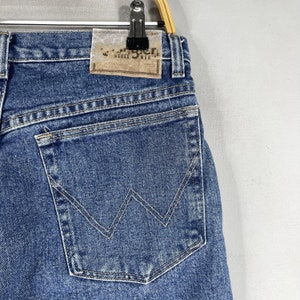 May include: A pair of blue denim jeans with a back pocket featuring a distinctive W-shaped stitching detail. The jeans have a brown leather patch with the text "Wrangler Rugged Wear" on the back waistband.