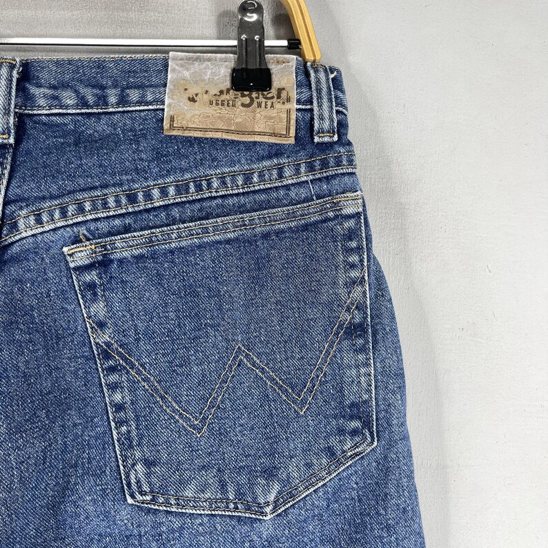 May include: A pair of blue denim jeans with a back pocket featuring a distinctive W-shaped stitching detail. The jeans have a brown leather patch with the text "Wrangler Rugged Wear" on the back waistband.