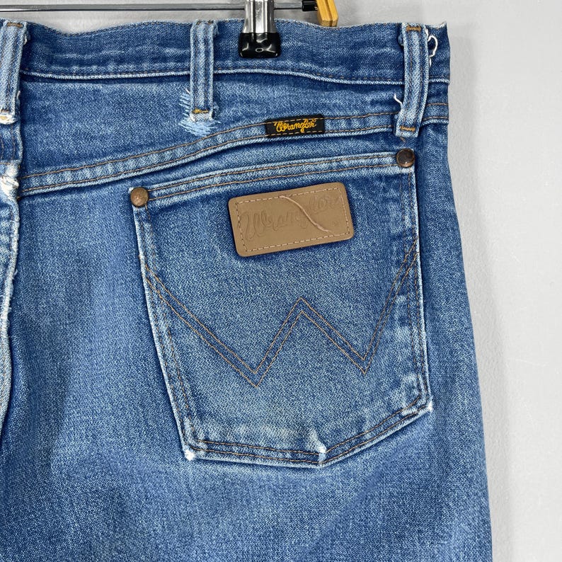 Pode incluir: Close-up de cal&ccedil;as de ganga azuis com um bolso traseiro com um design 'W' costurado. Uma etiqueta de couro castanho com o log&oacute;tipo 'Wrangler' &eacute; vis&iacute;vel. As cal&ccedil;as de ganga t&ecirc;m um estilo cl&aacute;ssico e vintage.
