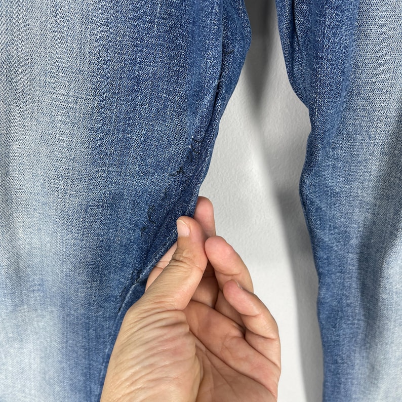 May include: Close-up of blue denim jeans. The jeans have a faded wash effect, with lighter shades at the thighs and knees. The fabric is held by a hand, showing the texture and stitching details. The background is a plain white wall.