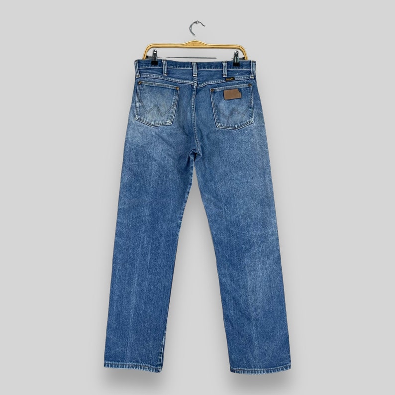 May include: A pair of blue denim jeans hanging on a wooden hanger. The jeans have a brown leather patch on the back pocket with the word "Wrangler" printed on it.