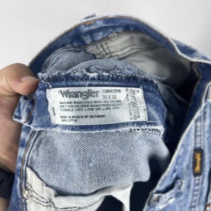 Pu&ograve; includere: Un primo piano dell'interno di un paio di jeans blu chiaro con un'etichetta Wrangler. L'etichetta dice "13MWZPW 32 X 32" e include le istruzioni di lavaggio. L'etichetta dice anche "Made in Mexico of US Fabric 100% Cotton."