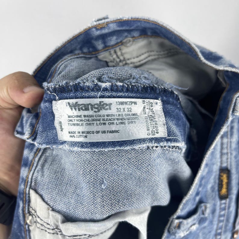 Pu&ograve; includere: Un primo piano dell'interno di un paio di jeans blu chiaro con un'etichetta Wrangler. L'etichetta dice "13MWZPW 32 X 32" e include le istruzioni di lavaggio. L'etichetta dice anche "Made in Mexico of US Fabric 100% Cotton."