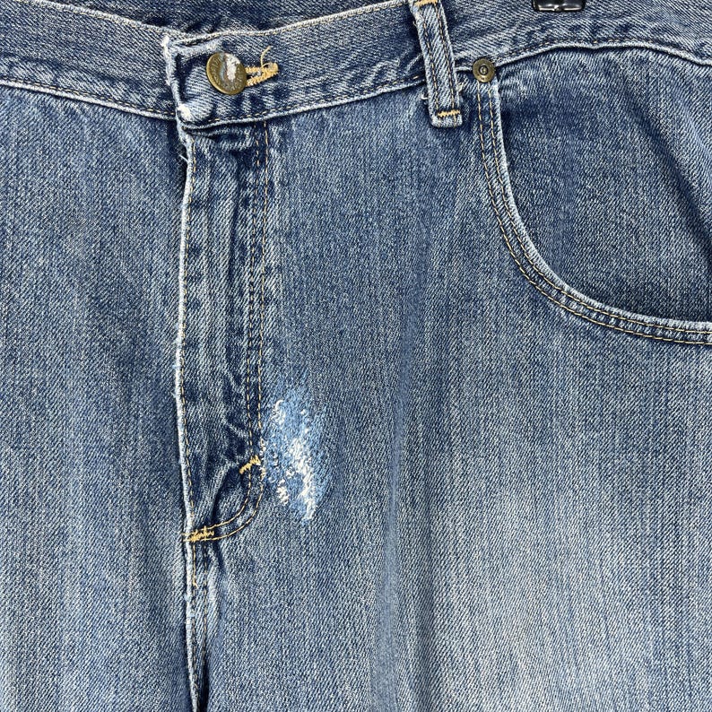 May include: Close-up of blue denim jeans. The jeans have a button closure, a coin pocket, and a distressed area on the front. The denim has a faded appearance.