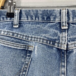May include: Close-up of the back pocket of a pair of light blue denim jeans. The pocket has a small tear in the fabric.