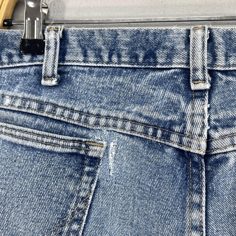 May include: Close-up of the back pocket of a pair of light blue denim jeans. The pocket has a small tear in the fabric.