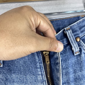May include: Close-up of a blue denim zipper fly with a gold zipper pull that says "Sunvil".