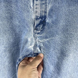 Pu&ograve; includere: Primo piano di un paio di jeans in denim blu chiaro con un'area rovinata sul davanti. I jeans hanno una cucitura al centro e un aspetto sbiadito.