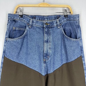 K&ouml;nnte beinhalten: Blaue Jeans aus Denim mit braunen Stoffbahnen am unteren Ende. Die Jeans haben einen Knopfverschluss und G&uuml;rtelschlaufen.