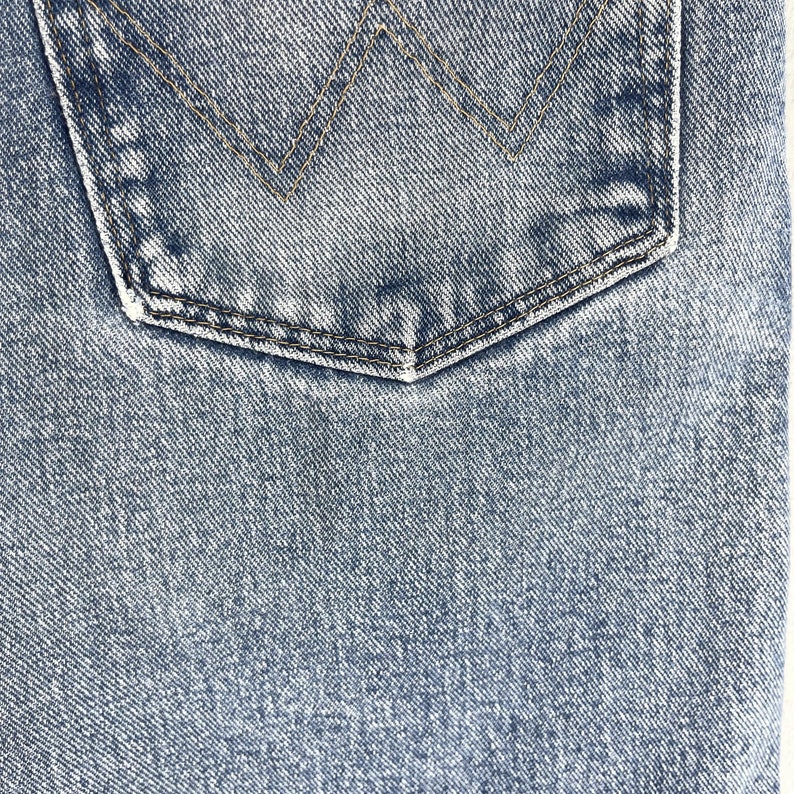 May include: Close-up of a blue denim fabric with a back pocket. The pocket has a stitched detail in the shape of a triangle.