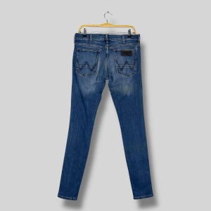 May include: Blue denim jeans with a classic five-pocket design, hanging on a yellow hanger. The back pockets feature a distinctive 'W' stitching. A brown leather patch is visible on the back waistband.