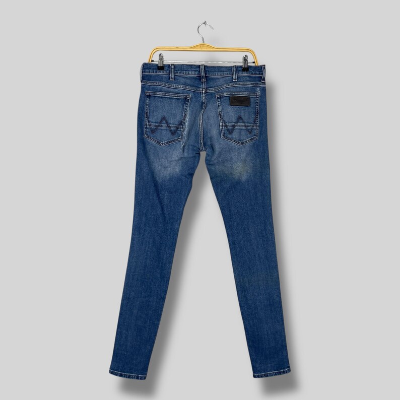 May include: Blue denim jeans with a classic five-pocket design, hanging on a yellow hanger. The back pockets feature a distinctive 'W' stitching. A brown leather patch is visible on the back waistband.