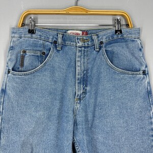 Puede incluir: Vaqueros de mezclilla azul claro con un dise&ntilde;o cl&aacute;sico de cinco bolsillos. Los vaqueros tienen cierre de bot&oacute;n y cremallera, trabillas para el cintur&oacute;n y la etiqueta de la marca "Wrangler". Los vaqueros est&aacute;n colgados de una percha de madera.
