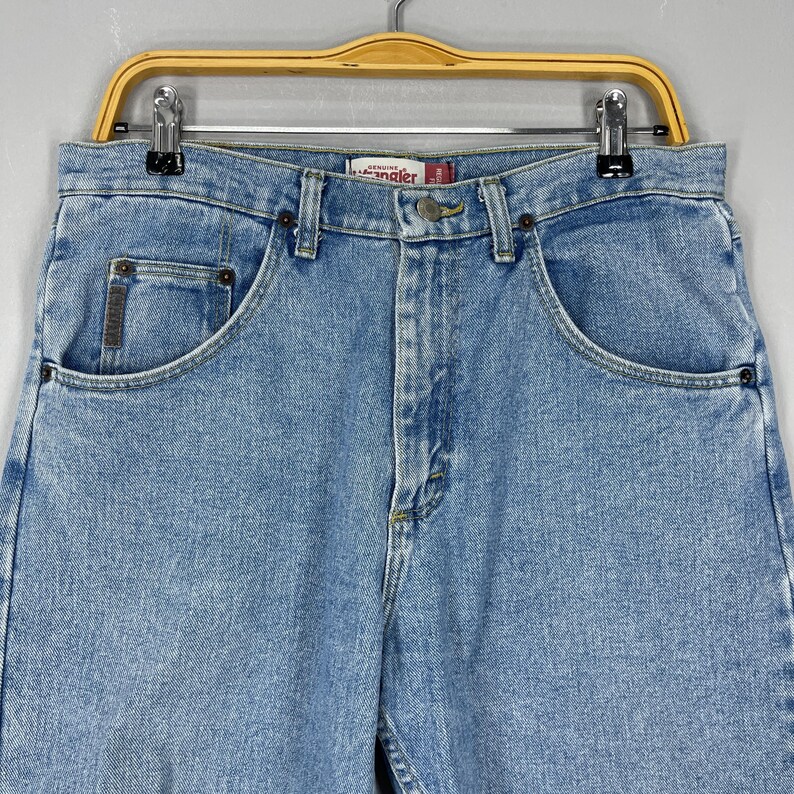 Puede incluir: Vaqueros de mezclilla azul claro con un dise&ntilde;o cl&aacute;sico de cinco bolsillos. Los vaqueros tienen cierre de bot&oacute;n y cremallera, trabillas para el cintur&oacute;n y la etiqueta de la marca "Wrangler". Los vaqueros est&aacute;n colgados de una percha de madera.