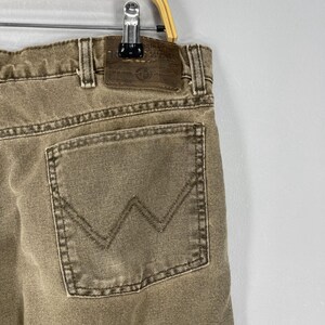 May include: Close-up of a pair of tan-colored denim jeans. The jeans feature a back pocket with a stitched "W" design and a leather patch. The jeans are hanging on a metal hanger.