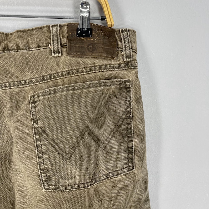May include: Close-up of a pair of tan-colored denim jeans. The jeans feature a back pocket with a stitched "W" design and a leather patch. The jeans are hanging on a metal hanger.
