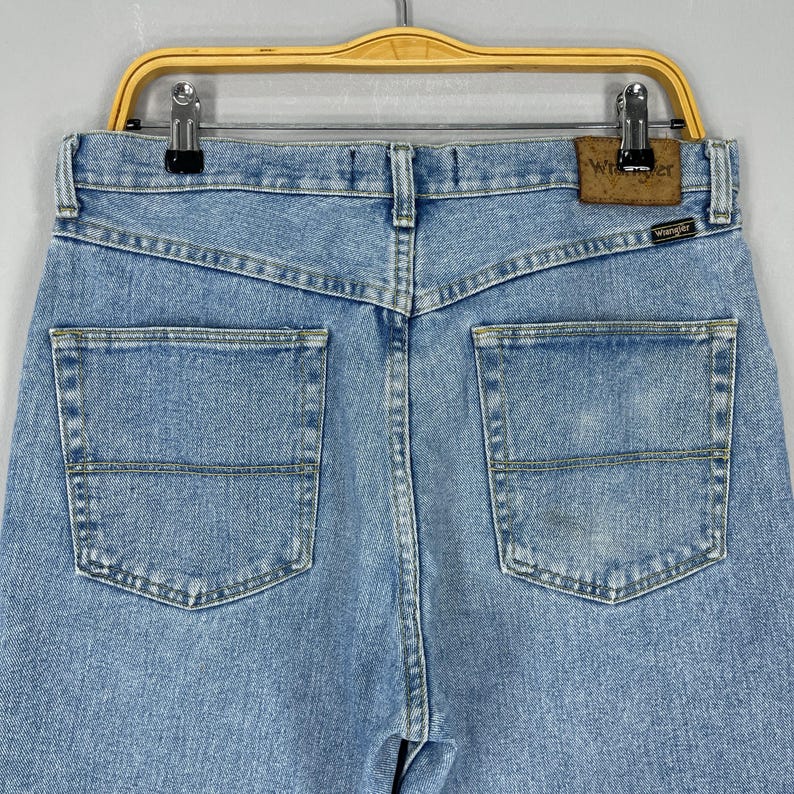 Puede incluir: Vaqueros de mezclilla azul claro con dos bolsillos traseros y un parche de la marca Wrangler. Los vaqueros est&aacute;n colgados de una percha de madera. La mezclilla tiene un aspecto descolorido.