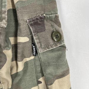 K&ouml;nnte beinhalten: Nahaufnahme von Camouflage-Stoff mit einer Tasche und Knopfdetails. Der Stoff weist eine Mischung aus Gr&uuml;n-, Braun- und Beiget&ouml;nen auf. Ein schwarzes Etikett mit dem Markennamen "AVIREX" ist sichtbar.
