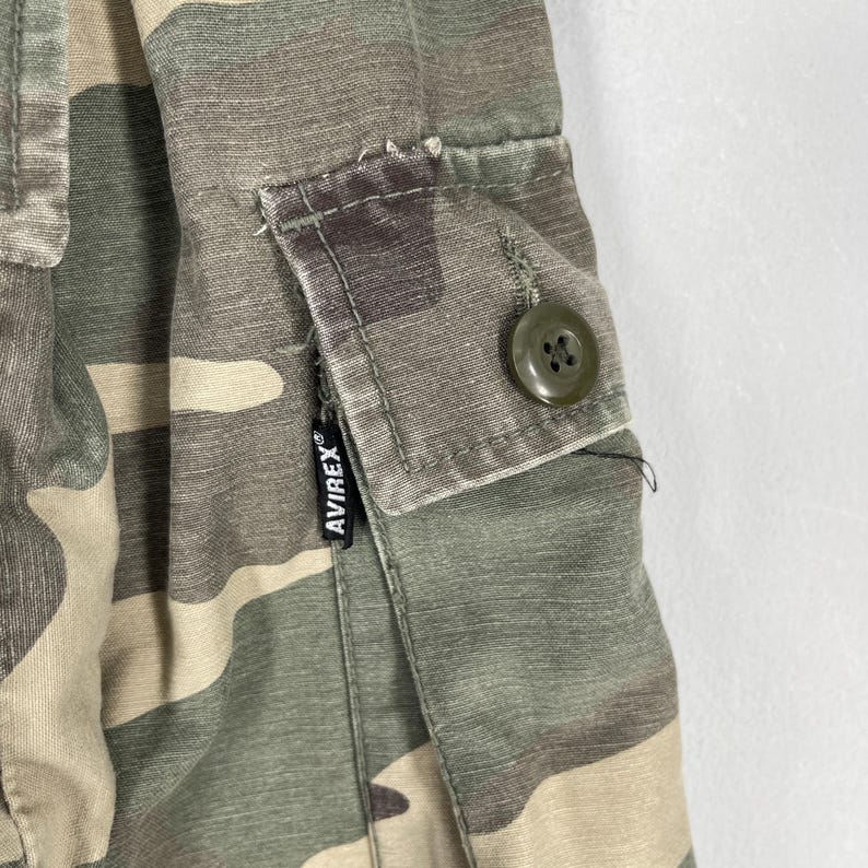 K&ouml;nnte beinhalten: Nahaufnahme von Camouflage-Stoff mit einer Tasche und Knopfdetails. Der Stoff weist eine Mischung aus Gr&uuml;n-, Braun- und Beiget&ouml;nen auf. Ein schwarzes Etikett mit dem Markennamen "AVIREX" ist sichtbar.