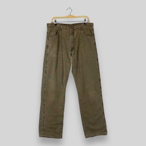 May include: Olive green denim jeans hanging on a gold-colored hanger. The jeans have a classic five-pocket design, a button and zipper closure, and a straight leg fit. The fabric appears worn and faded.