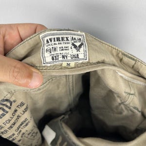 K&ouml;nnte beinhalten: Nahaufnahme des Inneren einer olivgr&uuml;nen Hose. Ein wei&szlig;es Etikett mit schwarzem Text lautet "AVIREX AL.5" und "037-NY-USA". Das Etikett enth&auml;lt auch den Text "SPEC.No. 66-7550" und "US PAT 2.06 1942". Die Gr&ouml;&szlig;e "M" ist unter dem Etikett aufgedruckt.