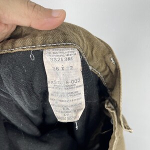 May include: Close-up of the inside of a pair of brown work pants. The tag reads "36 x 32" and includes other text. The pants are made of a durable material, with a black lining. The stitching is visible.