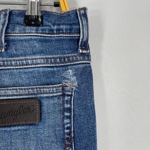 May include: Close-up of blue denim jeans with a distressed detail near the pocket. The jeans have a brown leather patch with the word "angler" on it. The jeans are hanging on a metal hanger.
