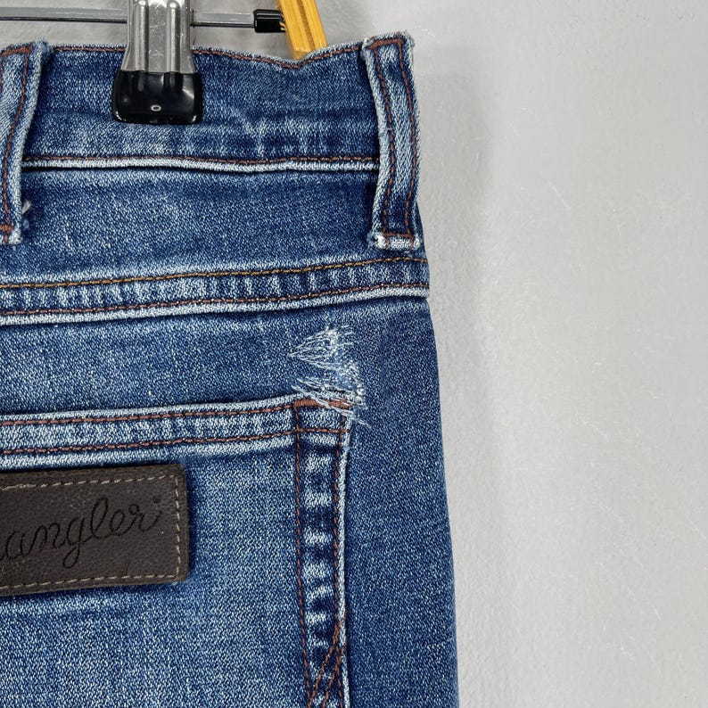 May include: Close-up of blue denim jeans with a distressed detail near the pocket. The jeans have a brown leather patch with the word "angler" on it. The jeans are hanging on a metal hanger.
